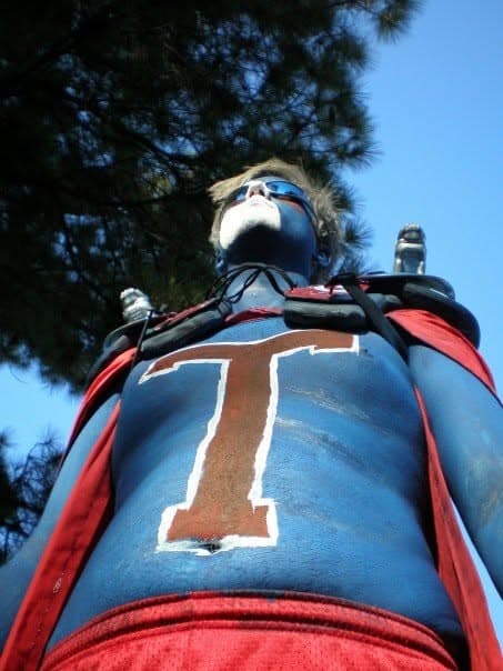 Bret painted blue with a large T on his chest, wearing shoulder pads beneath a bright sky